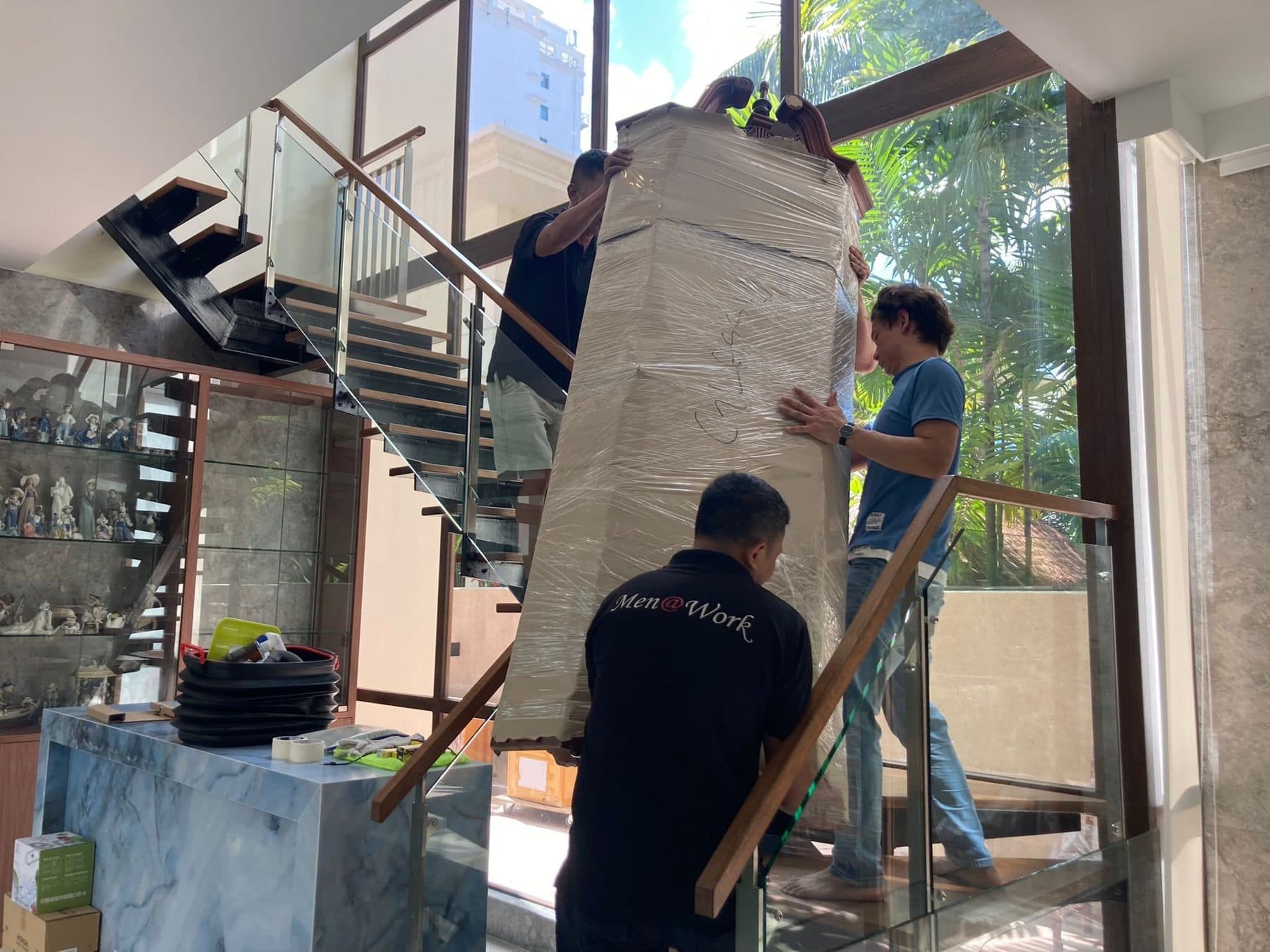 Three movers guiding a tall wrapped armoire on a staircase