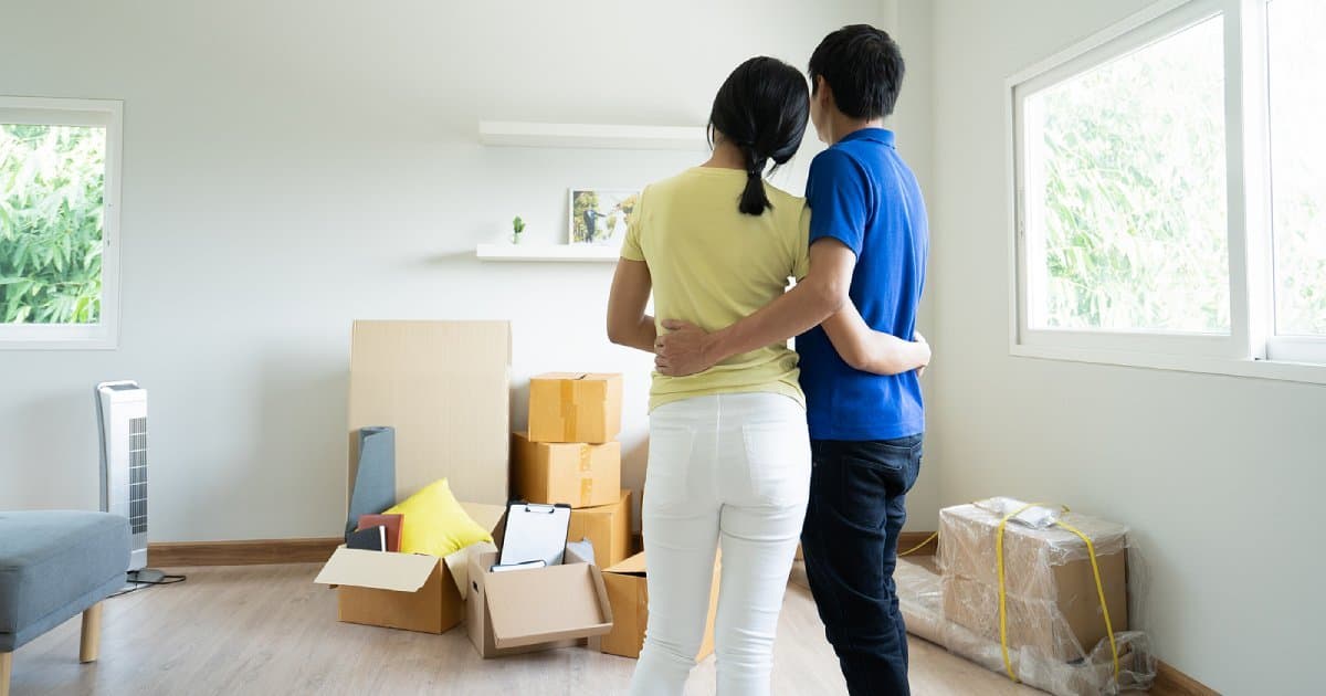 A couple with their arms around each other, looking at their new home among moving boxes