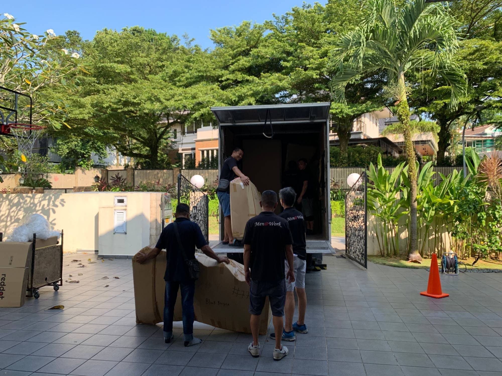 Reddot movers carrying a large boxed item to a truck in the driveway of a landed house