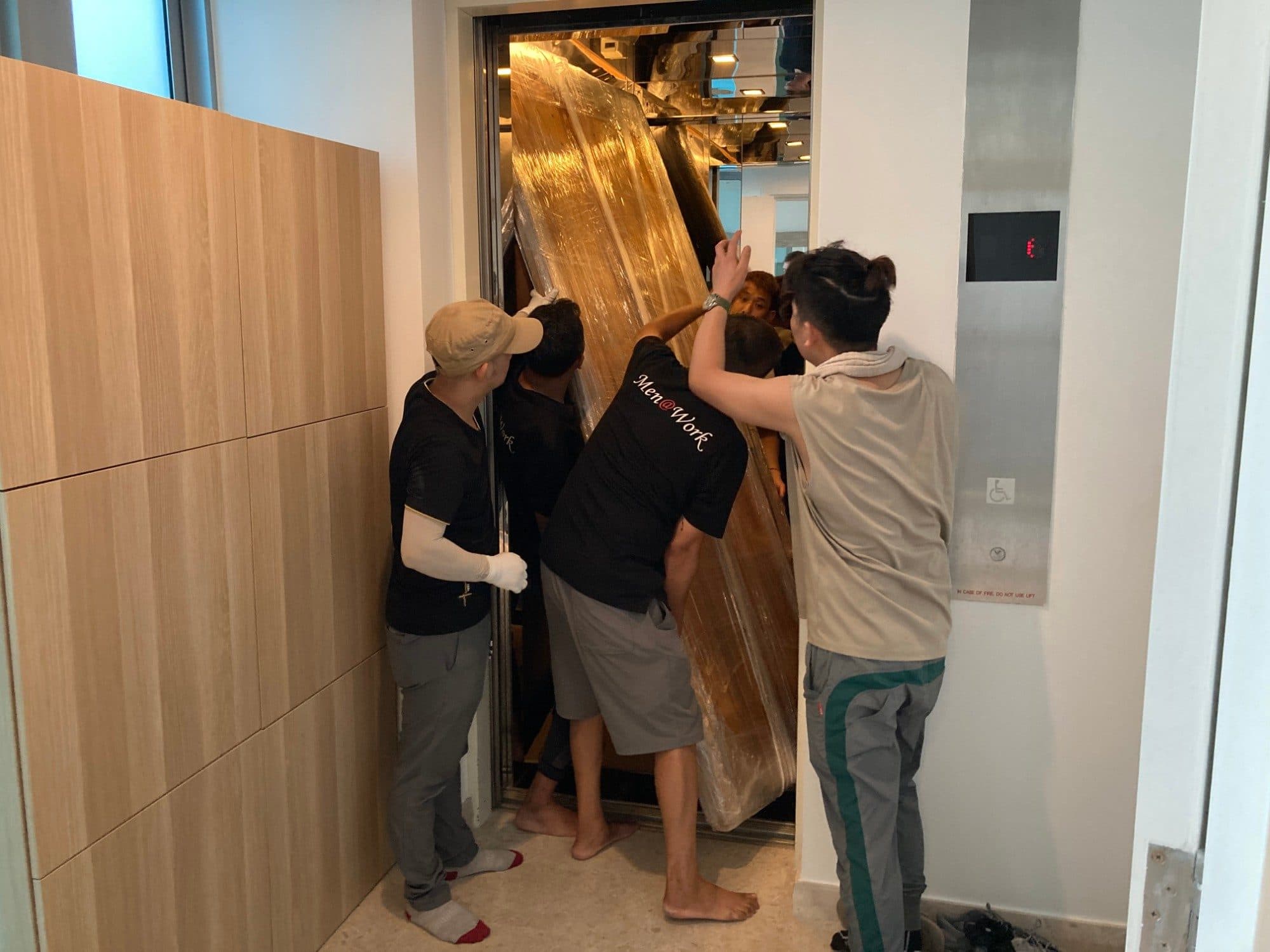 Movers easing a tall plastic-wrapped wooden piece into a lift