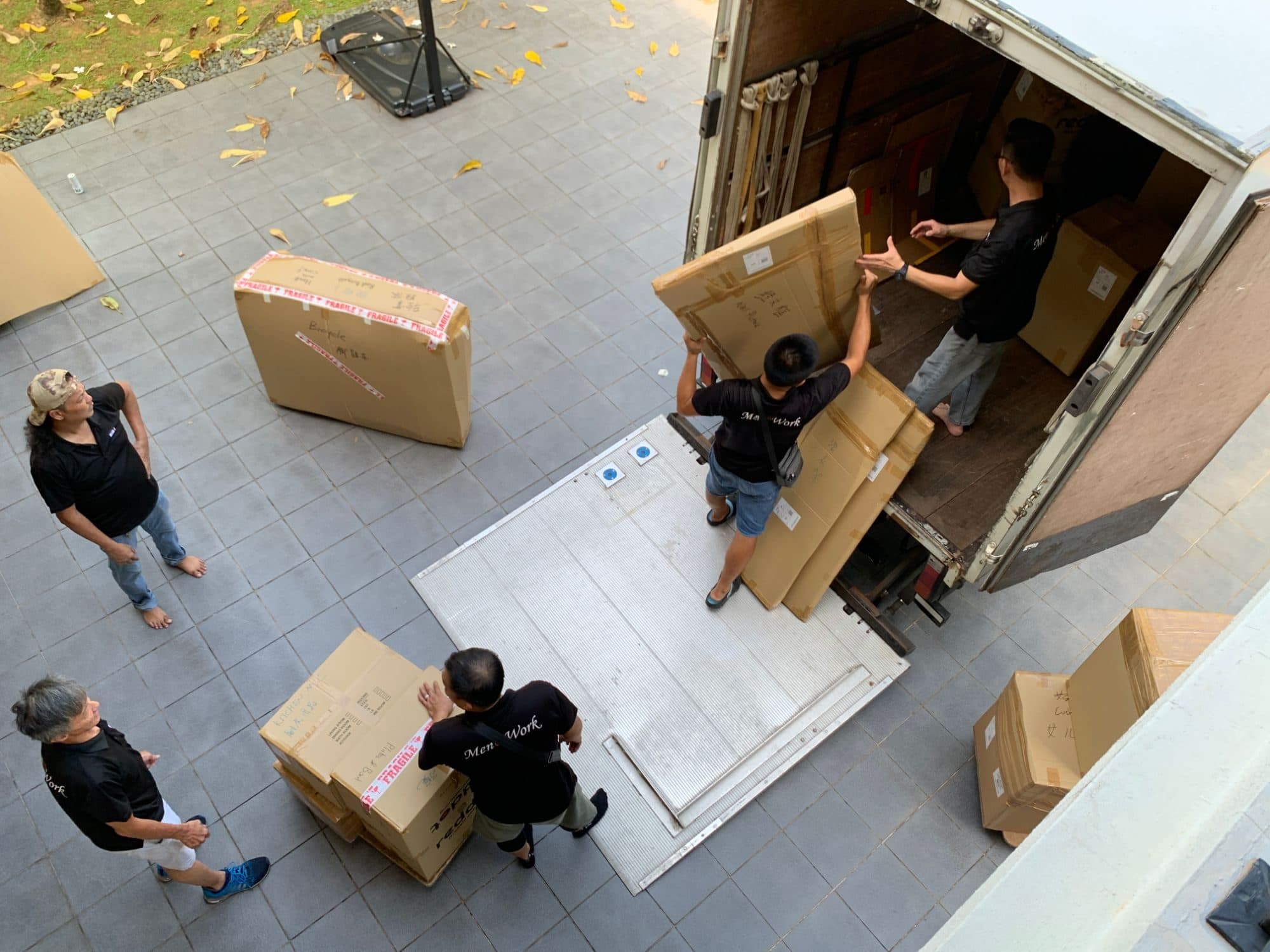 Reddot movers loading packed boxes and a boxed bicycle onto a truck's tail-lift, seen from above