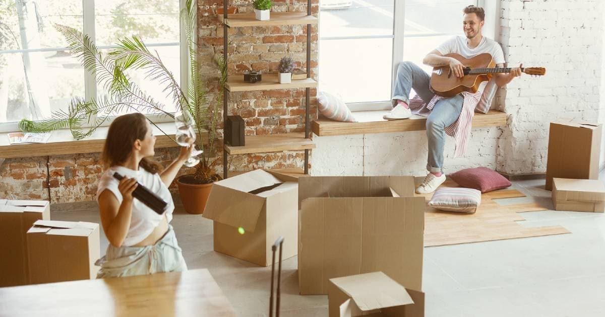 A woman holding a wine bottle and glass while a man plays guitar in a new flat full of moving boxes