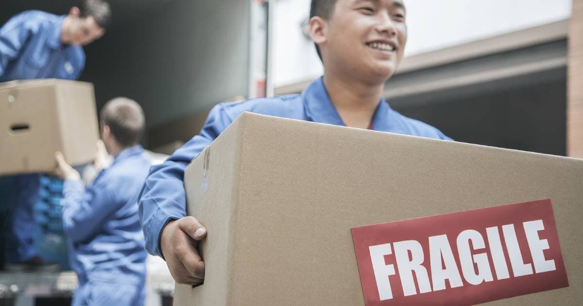 A smiling mover carrying a box marked “Fragile”, with colleagues unloading a truck behind him
