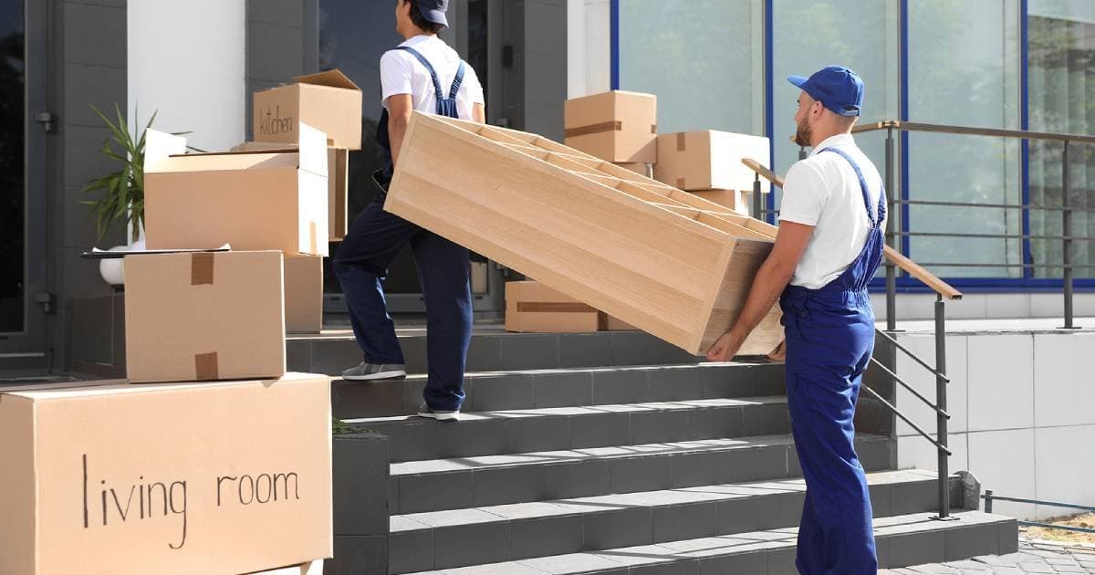 Two movers in overalls carrying furniture up the steps, past boxes labelled by room